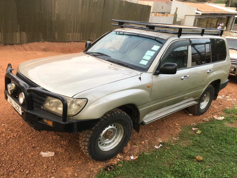 Land-cruiser-gx-hire-uganda - Rent 4x4 Car Uganda Beige 4x4 SUV with roof rack and front bull bar parked on a dirt lot, viewed from the front-left.