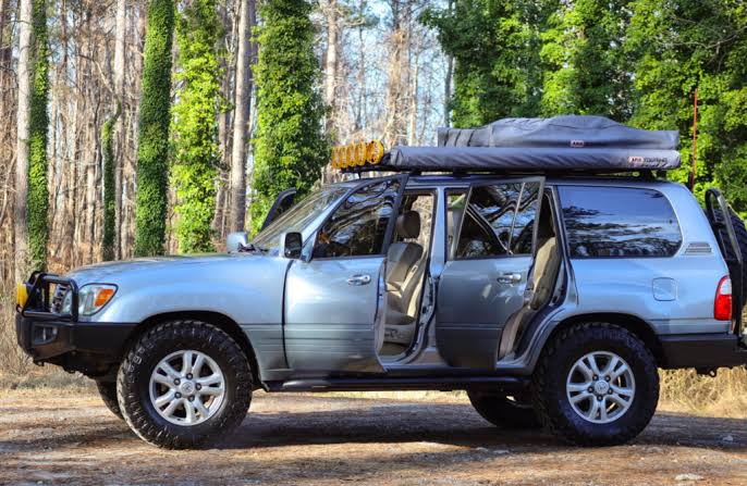 Blue SUV with roof rack and camping gear on top, doors open in a forest campsite.